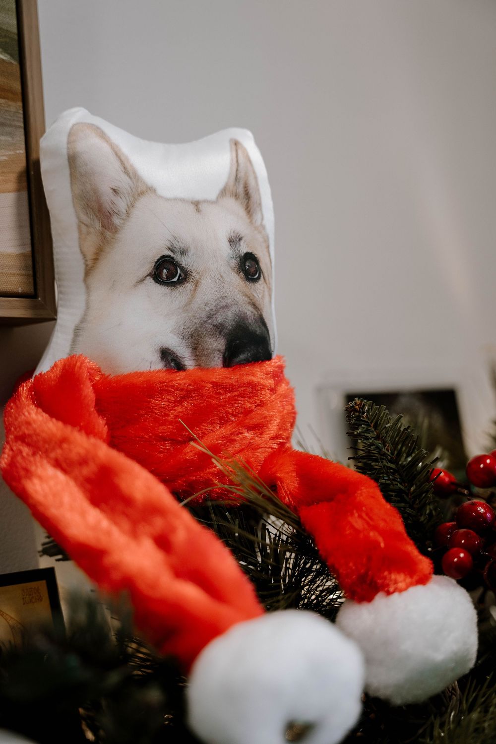A decorative dog face pillow wearing a red Santa hat and scarf nestled in a Christmas wreath with holiday berries.