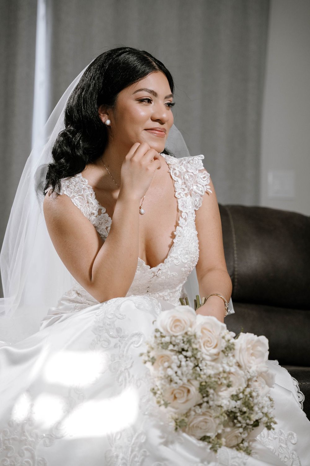 Elegant bride in lace wedding dress and veil sits holding white rose bouquet indoors.