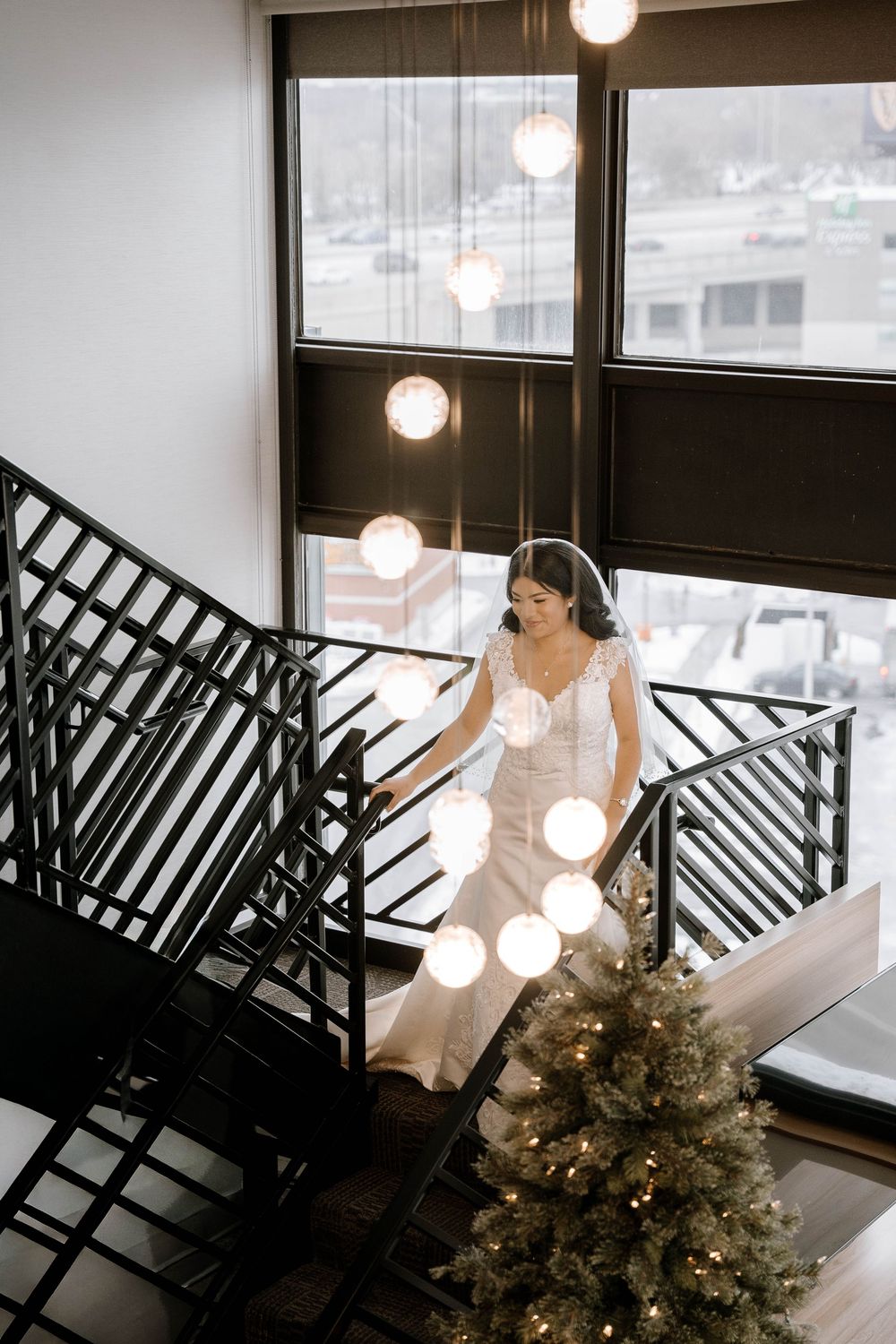 Bride in white gown descending elegant staircase with globe pendant lights and Christmas tree at modern wedding venue.