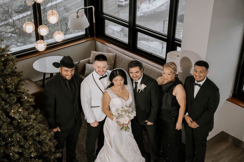 Wedding party of six poses indoors near Christmas tree and large windows in elegant black and white attire.