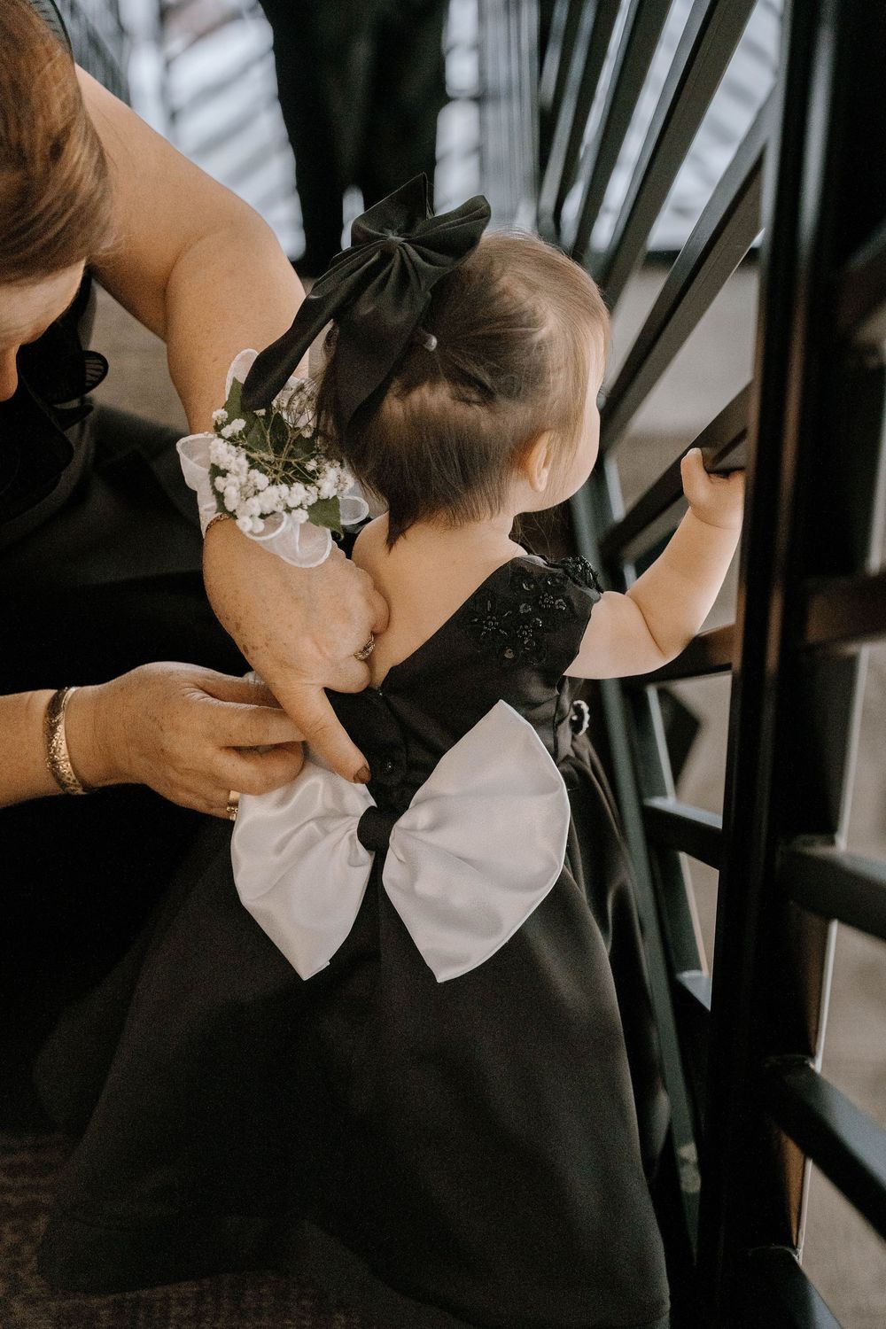 Baby girl in black dress with white bow and matching hair bow held by adult on staircase.