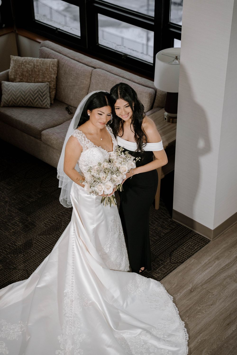 A bride in a white satin gown with veil holds a bouquet beside a bridesmaid in a black dress in a hotel suite.