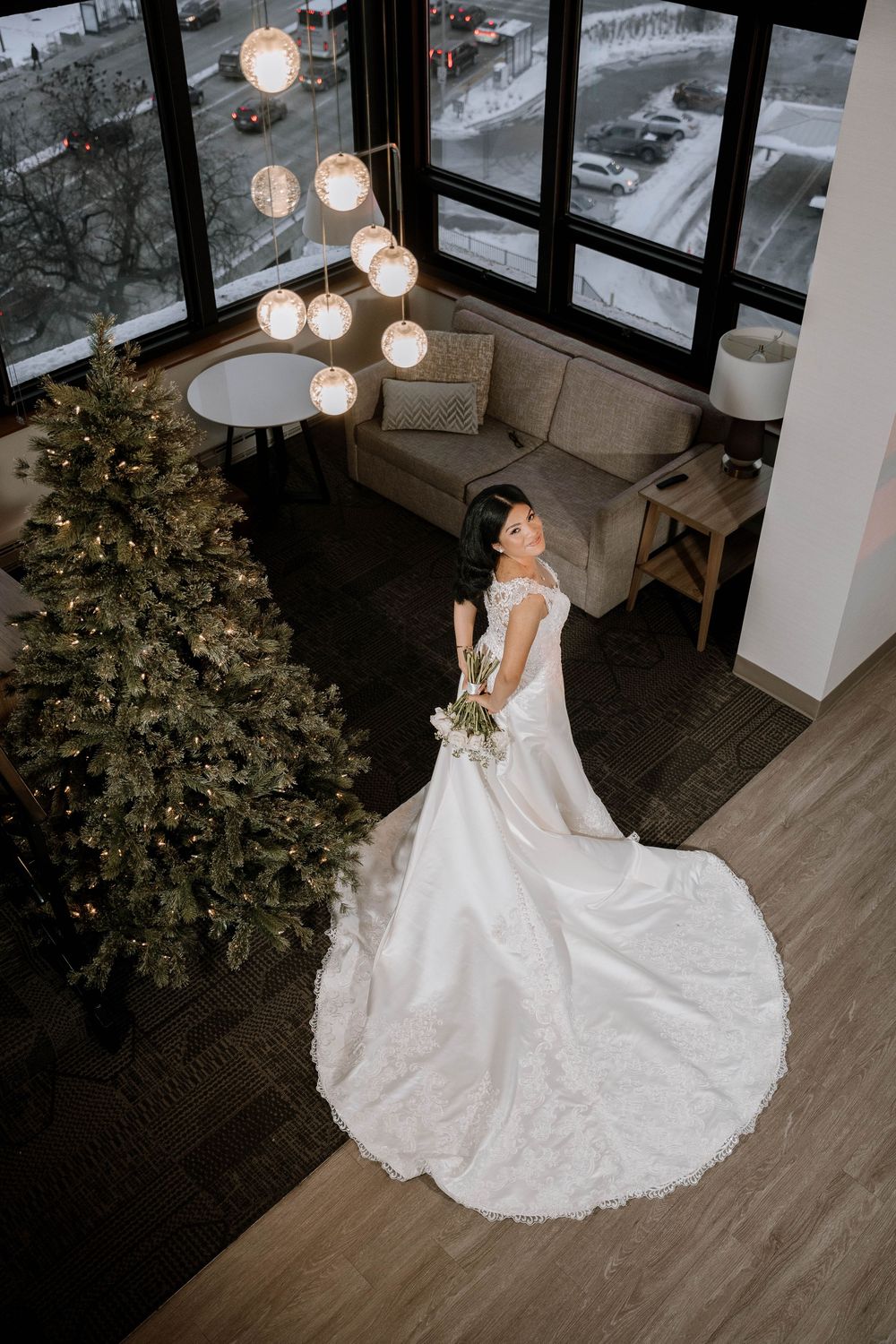 Aerial view of a bride in a white lace ball gown standing near a decorated Christmas tree indoors.