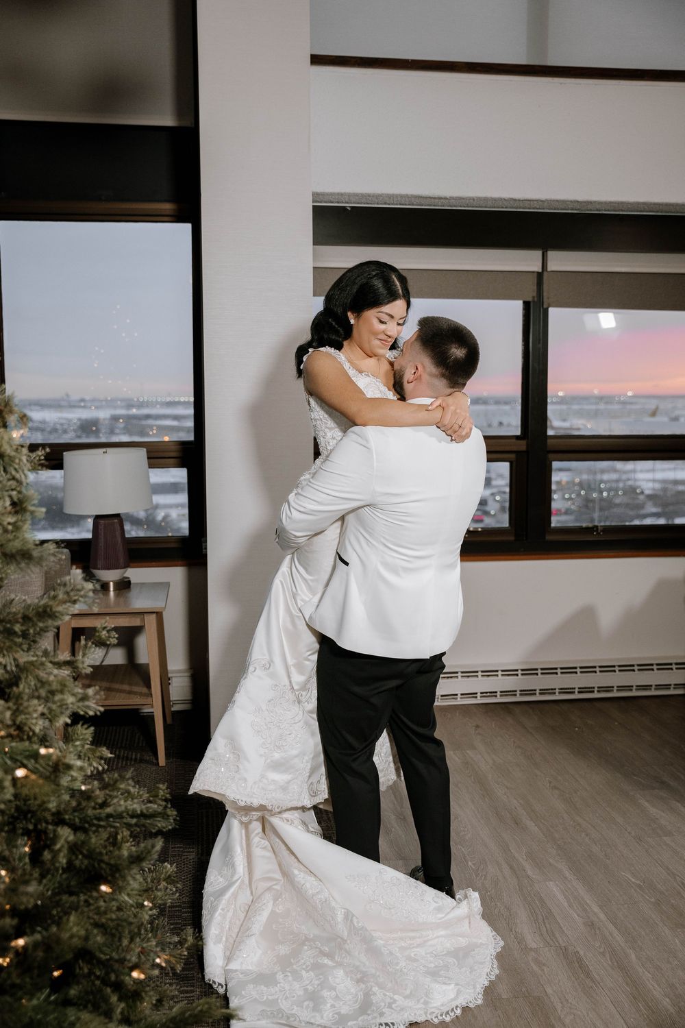 A groom in a white jacket lifts his bride in a flowing white gown near a Christmas tree at sunset.