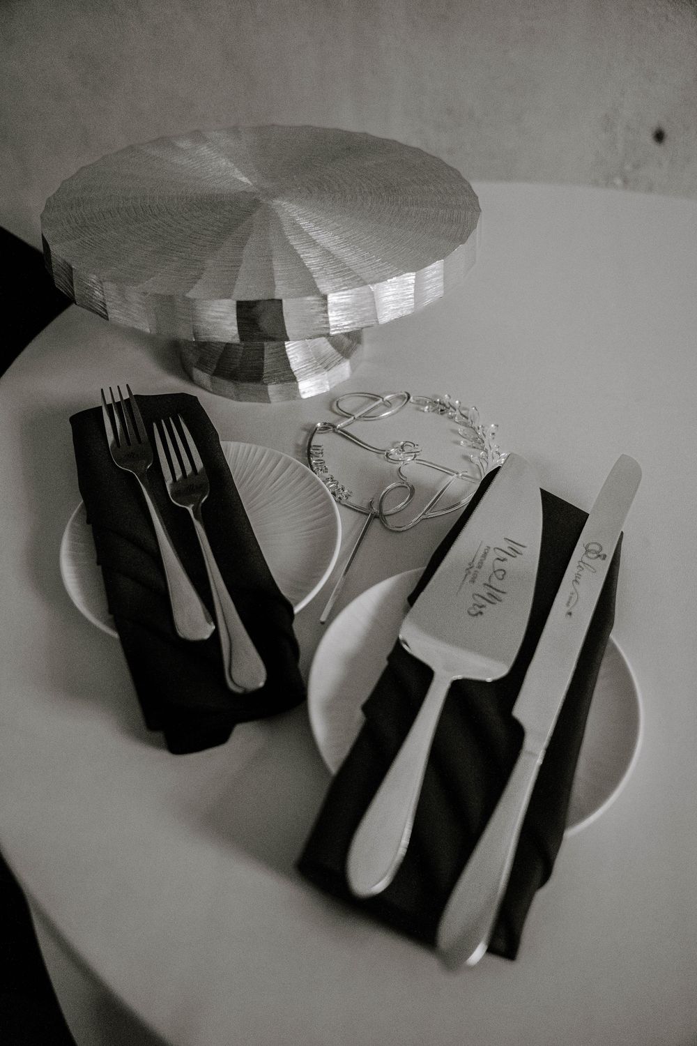 Black and white photo of wedding cake cutting set with engraved knife and server on white plates with black napkins.