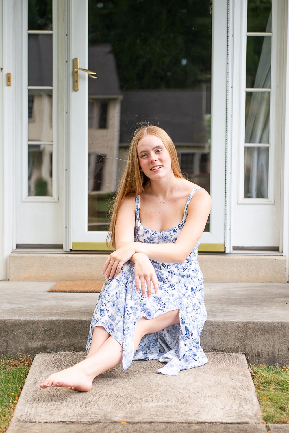 Someone in a blue and white floral dress poses casually on concrete steps outside a white building.
