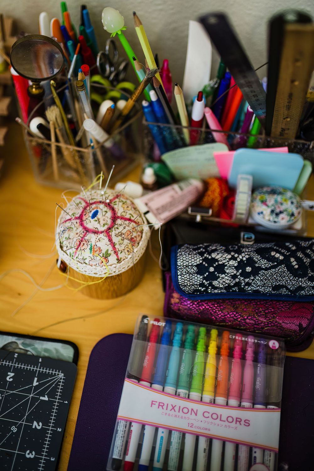 Colorful art supplies including markers, pens and craft materials arranged on a wooden desk with a pin cushion.