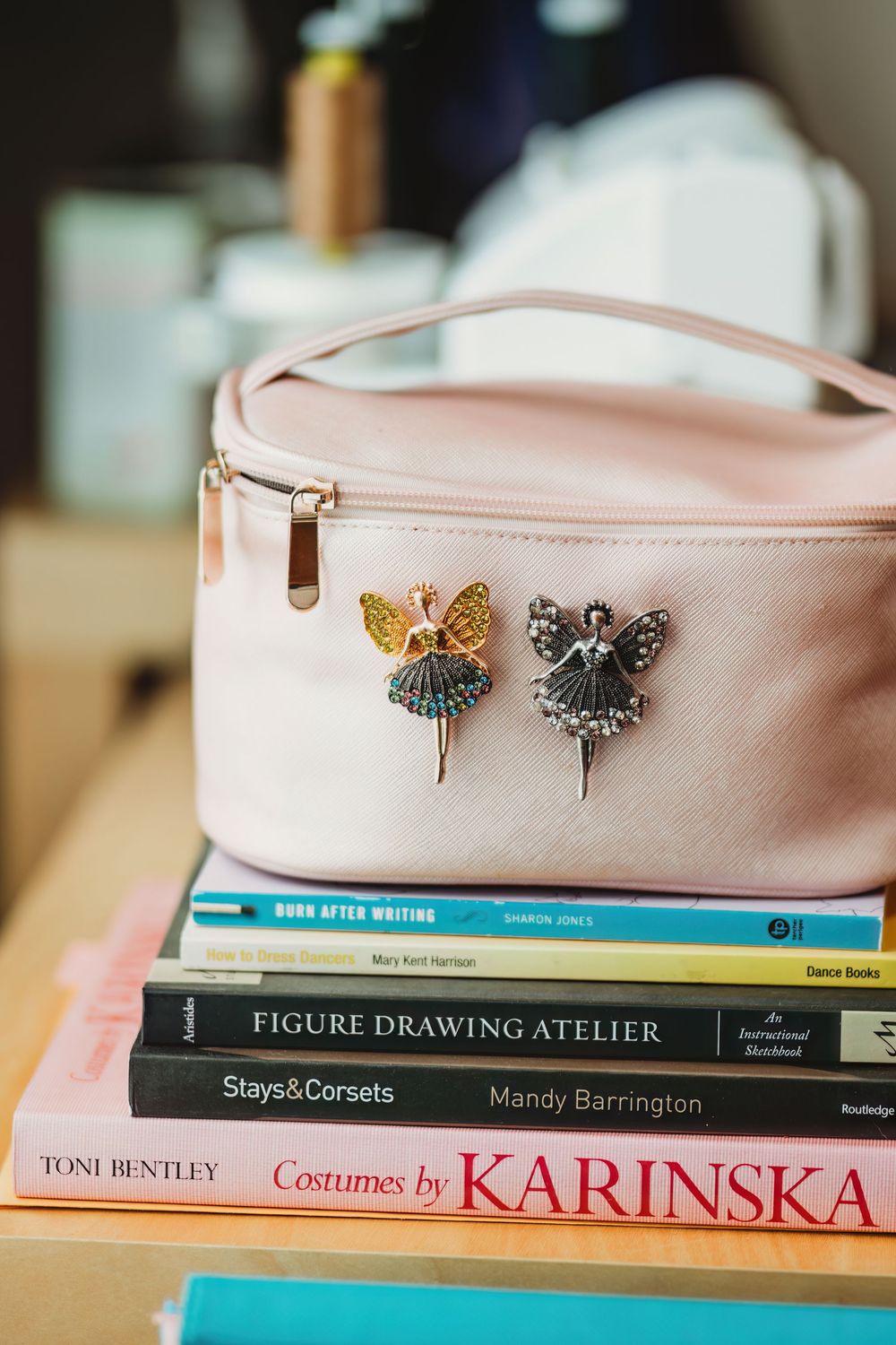 A pink leather bag sits atop a stack of art and fashion books including 'Figure Drawing Atelier' and 'Costumes by Karinska'.