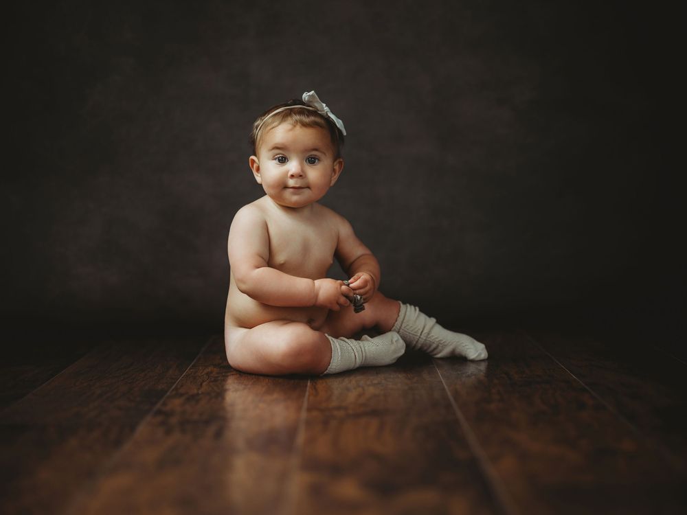 A series of artistic baby portraits against dark backgrounds showing developmental milestones on a wooden floor.