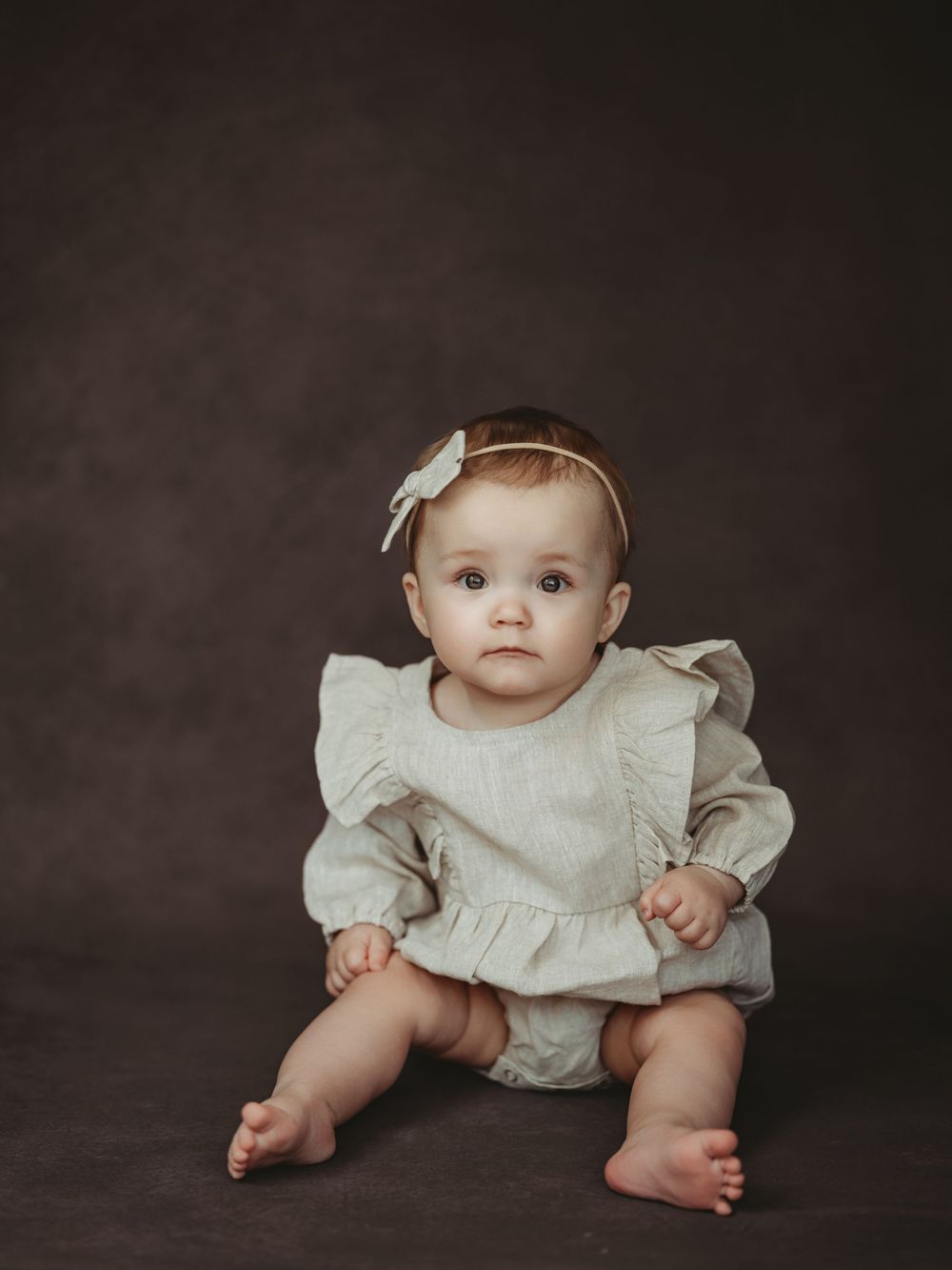 A baby wearing a white ruffled dress sits on a dark backdrop in a professional studio portrait.