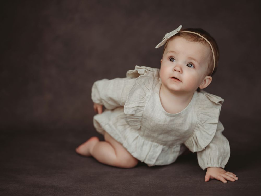 Baby in white ruffled dress lies on dark backdrop during professional photo session.