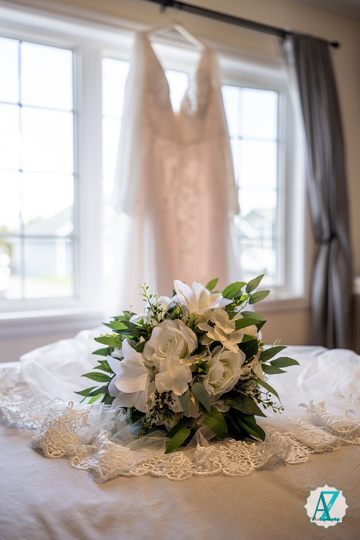 White bridal bouquet with roses and greenery rests on delicate lace fabric near sunlit windows.