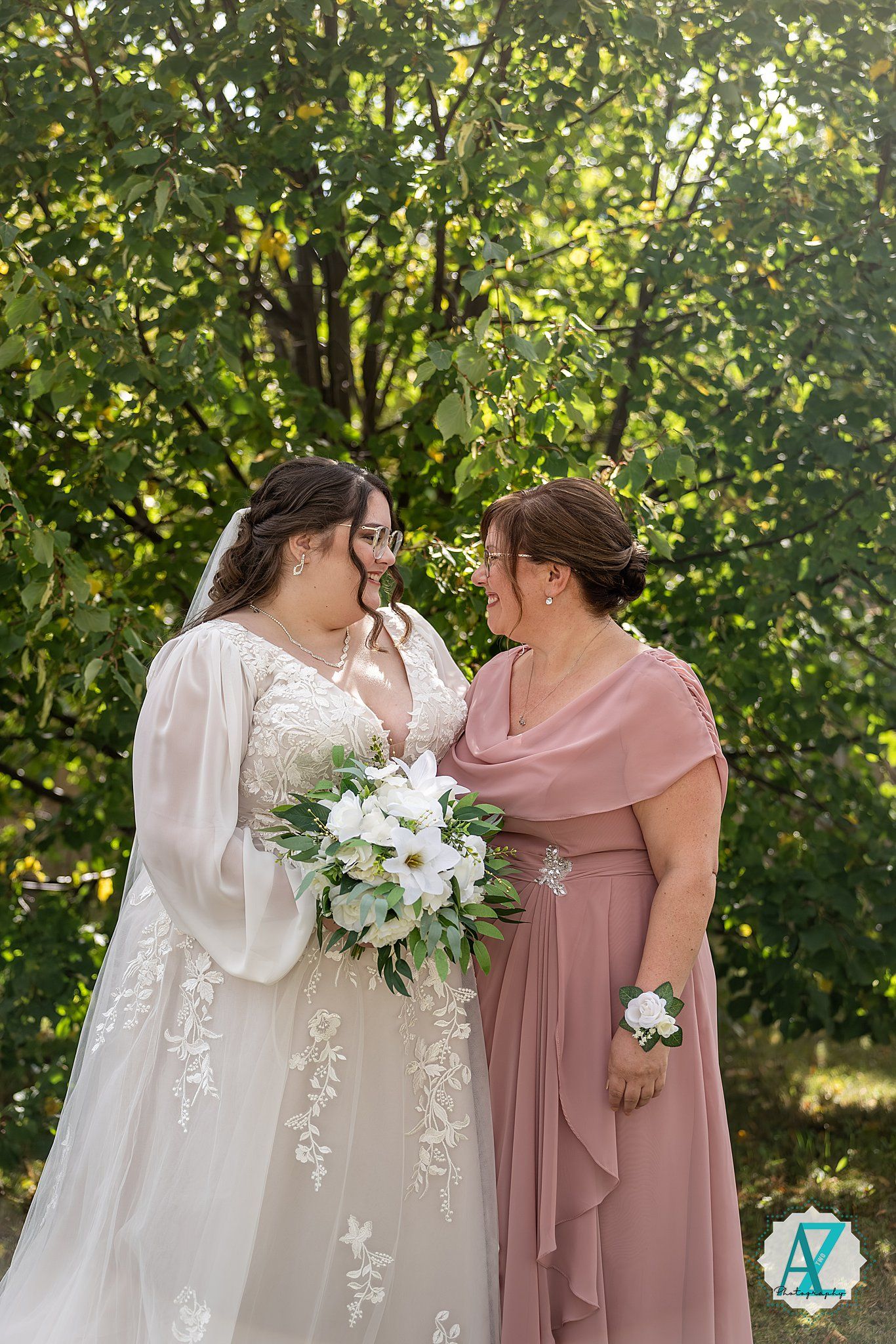 A bride in a white lace gown holds a white bouquet while sharing a special moment with someone in a mauve dress.