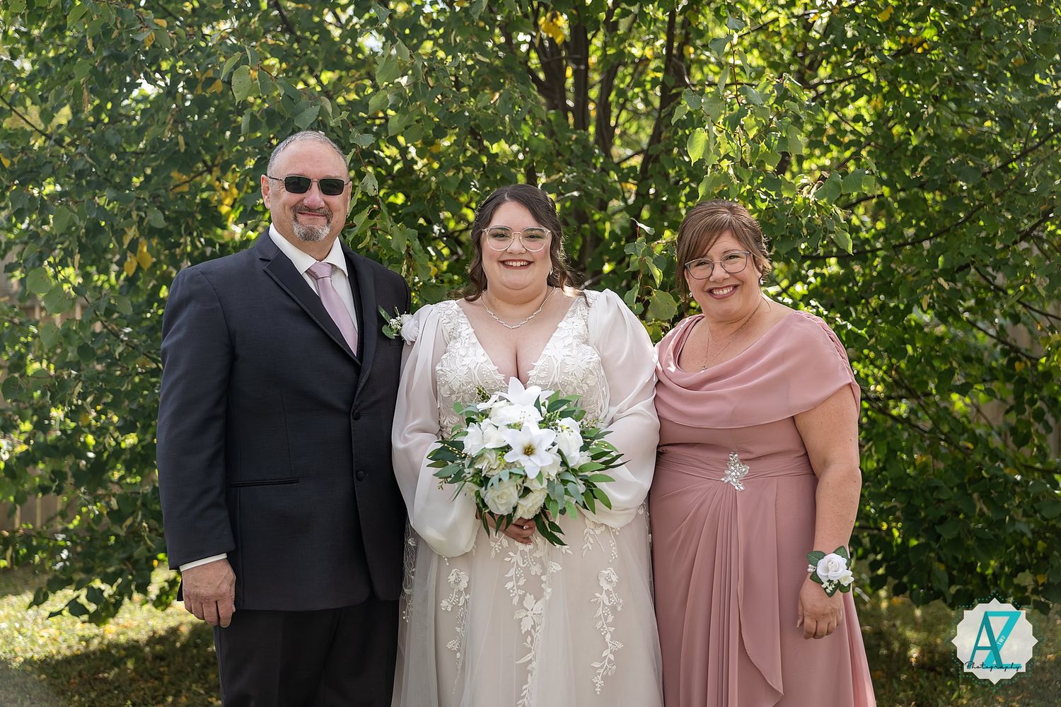 Wedding party poses outdoors in formal attire with pink bridesmaid dresses against a lush green garden backdrop.