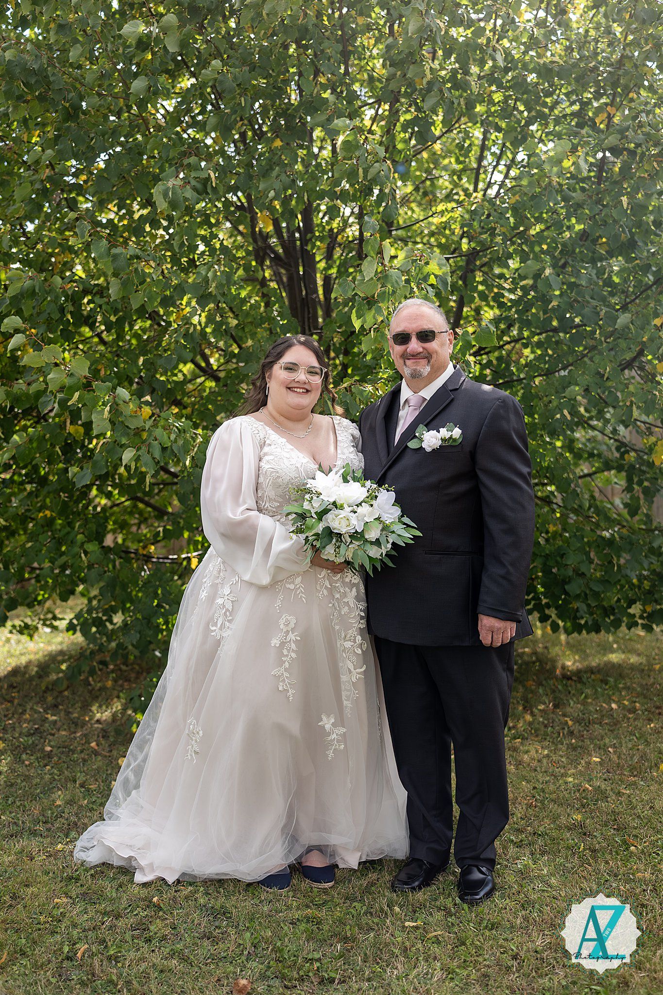 A wedding couple poses together against a leafy green backdrop while holding a white floral bouquet.