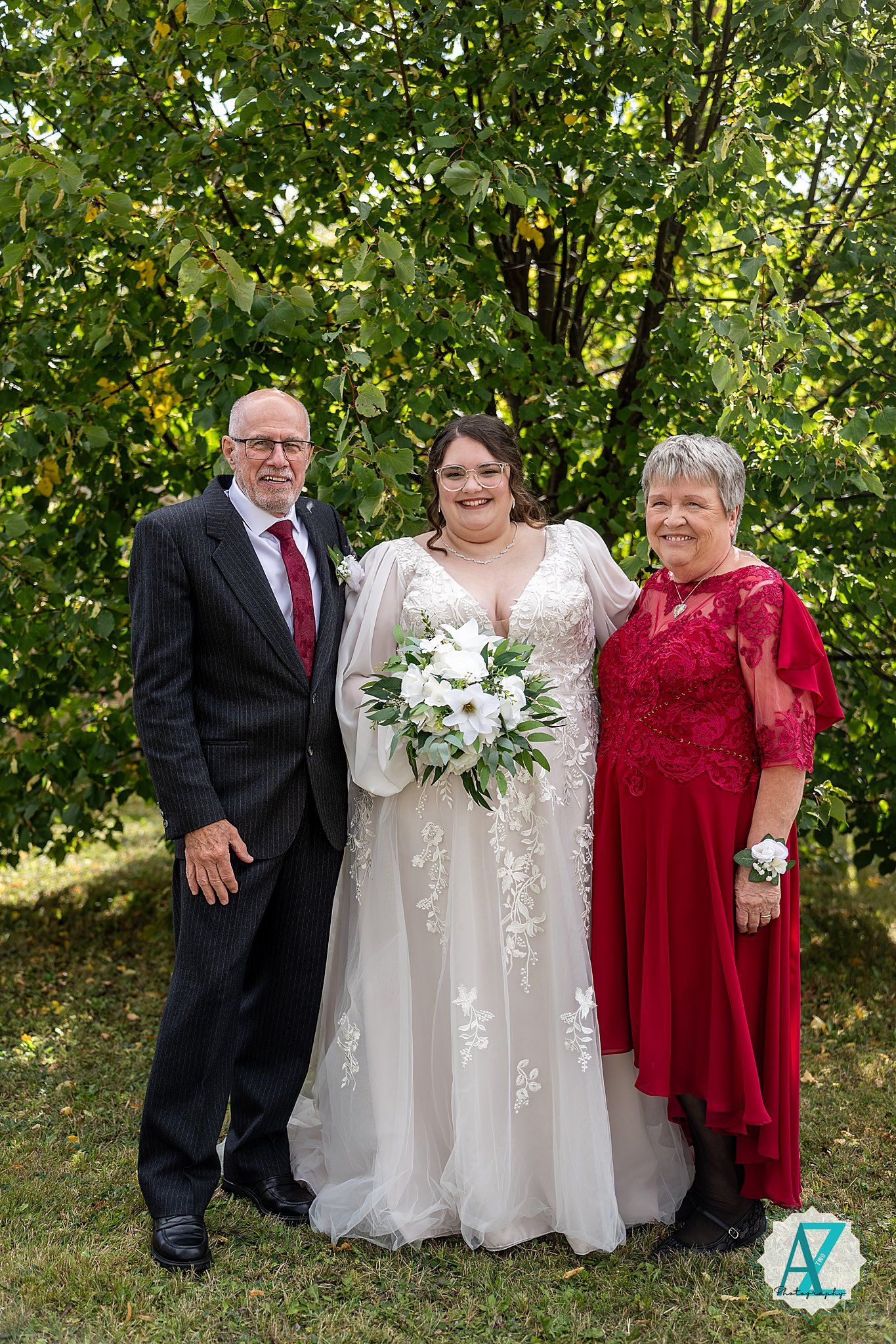 A wedding party poses outdoors in formal attire with the bride in white and guests in red dresses against a green backdrop.