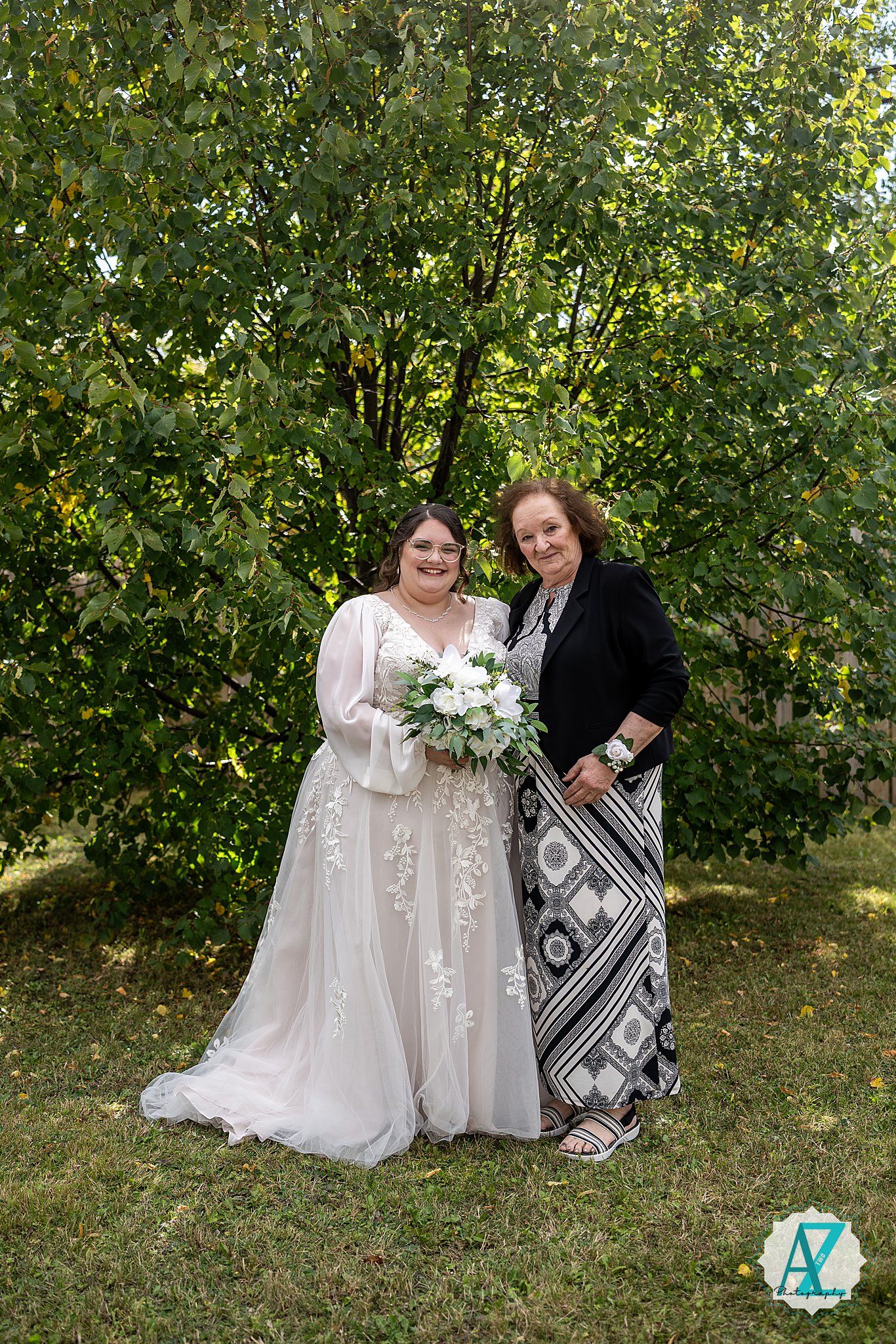 Two people in white dresses sit together on grass with trees in the background, holding a bouquet of white flowers.