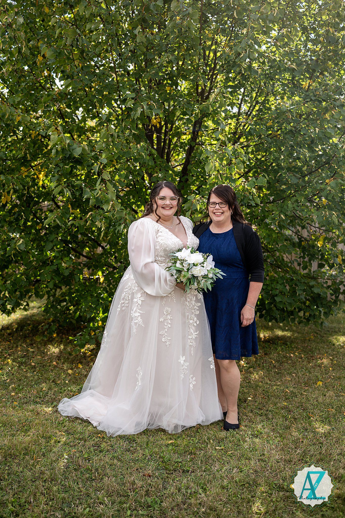 Two people stand together outdoors against a green leafy backdrop, one wearing a flowing white gown and one in navy blue.
