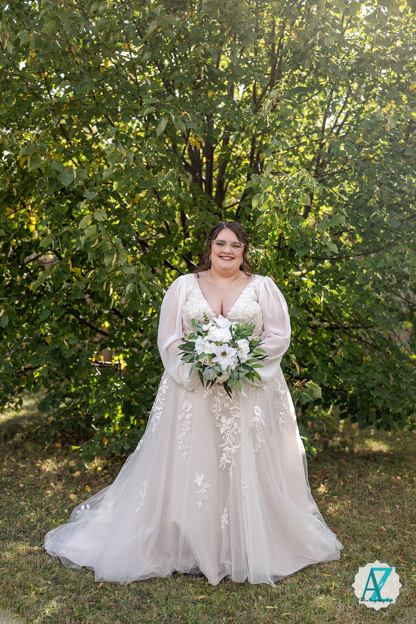 A bride in a flowing white lace wedding gown poses outdoors among green foliage holding a white floral bouquet.