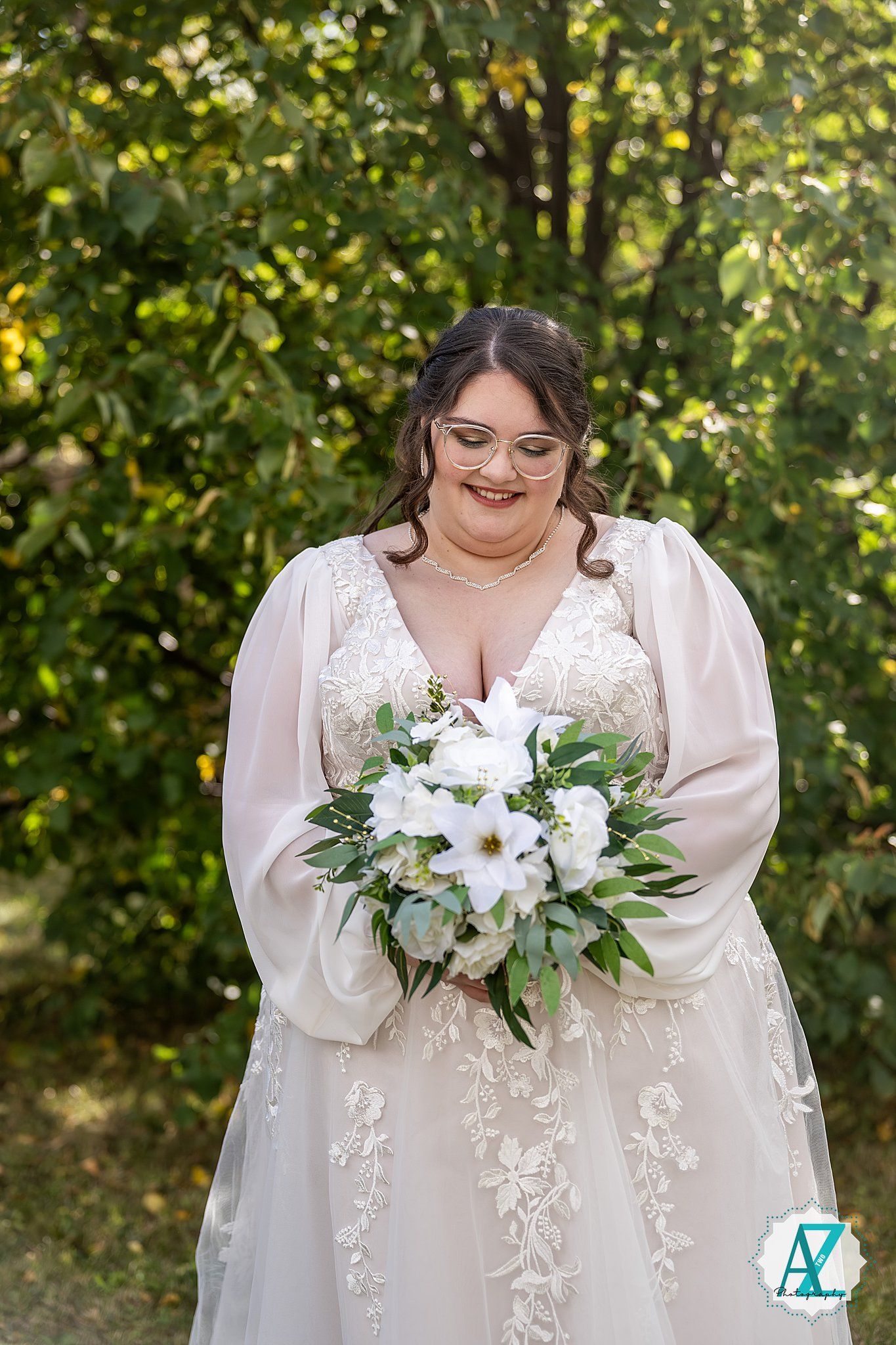 A bride in a flowing white gown holds a bouquet of white flowers and greenery against a sunlit garden backdrop.