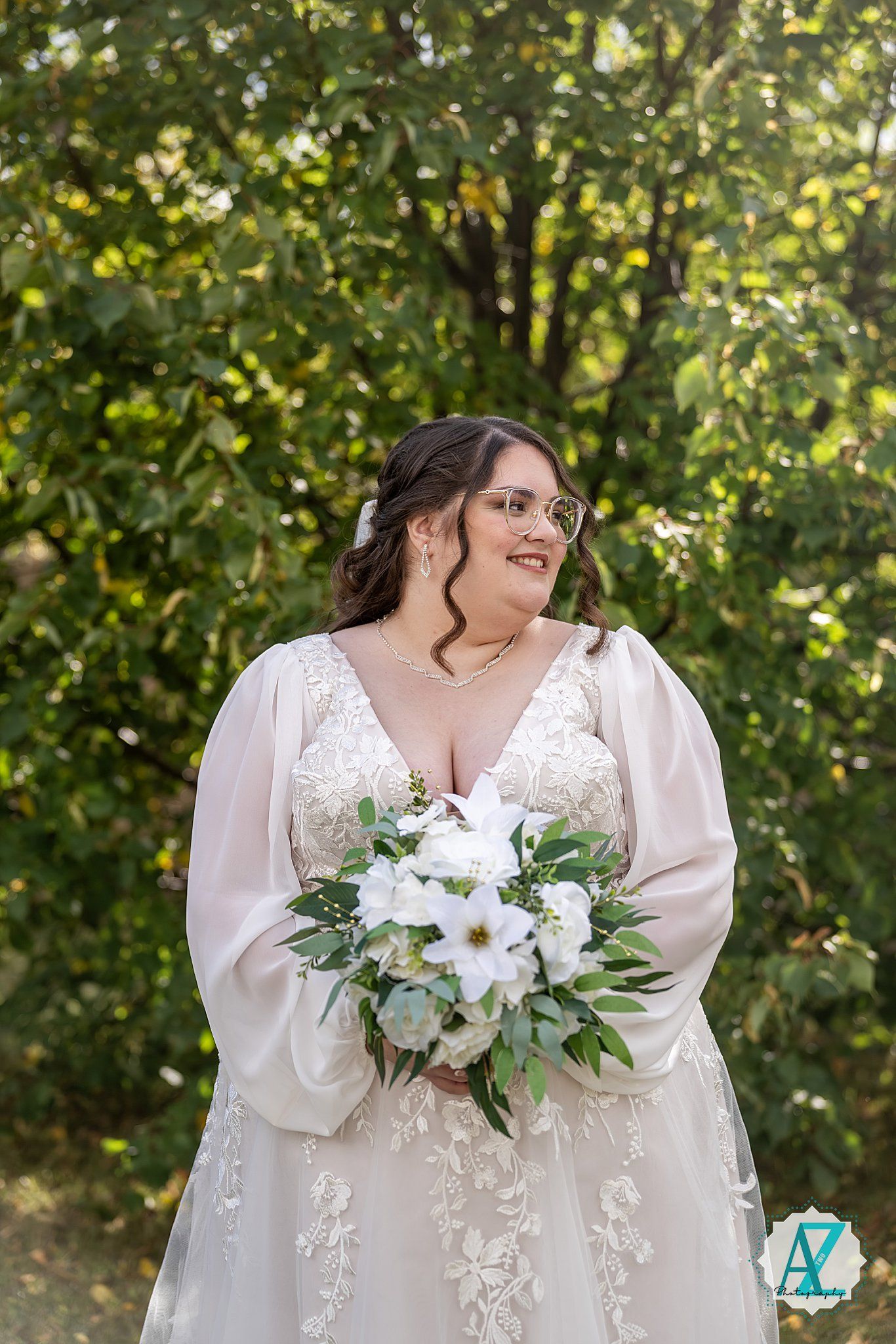 A beautiful wedding bouquet with white flowers and greenery is held against a lace dress with flowing sleeves in a garden setting.