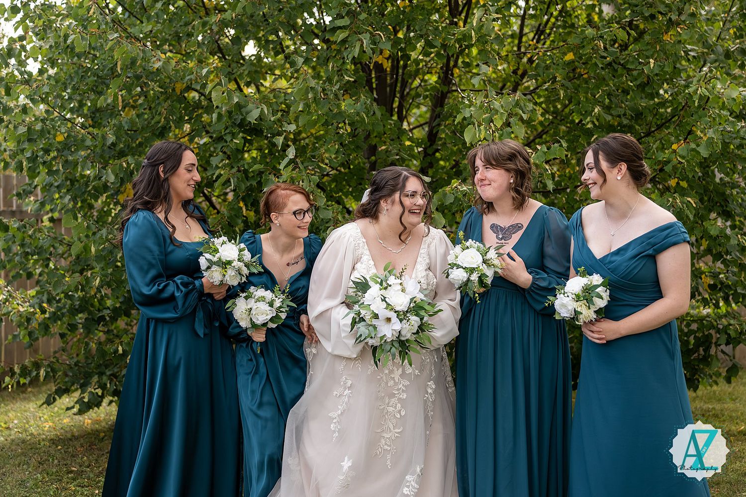 Bridesmaids in elegant teal dresses stand with brides holding white bouquets in a sunlit outdoor setting.