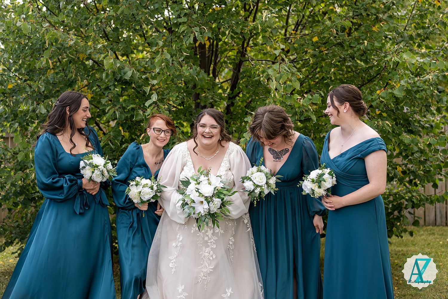 A bridal party wearing elegant teal blue dresses and holding white floral bouquets poses together outdoors.