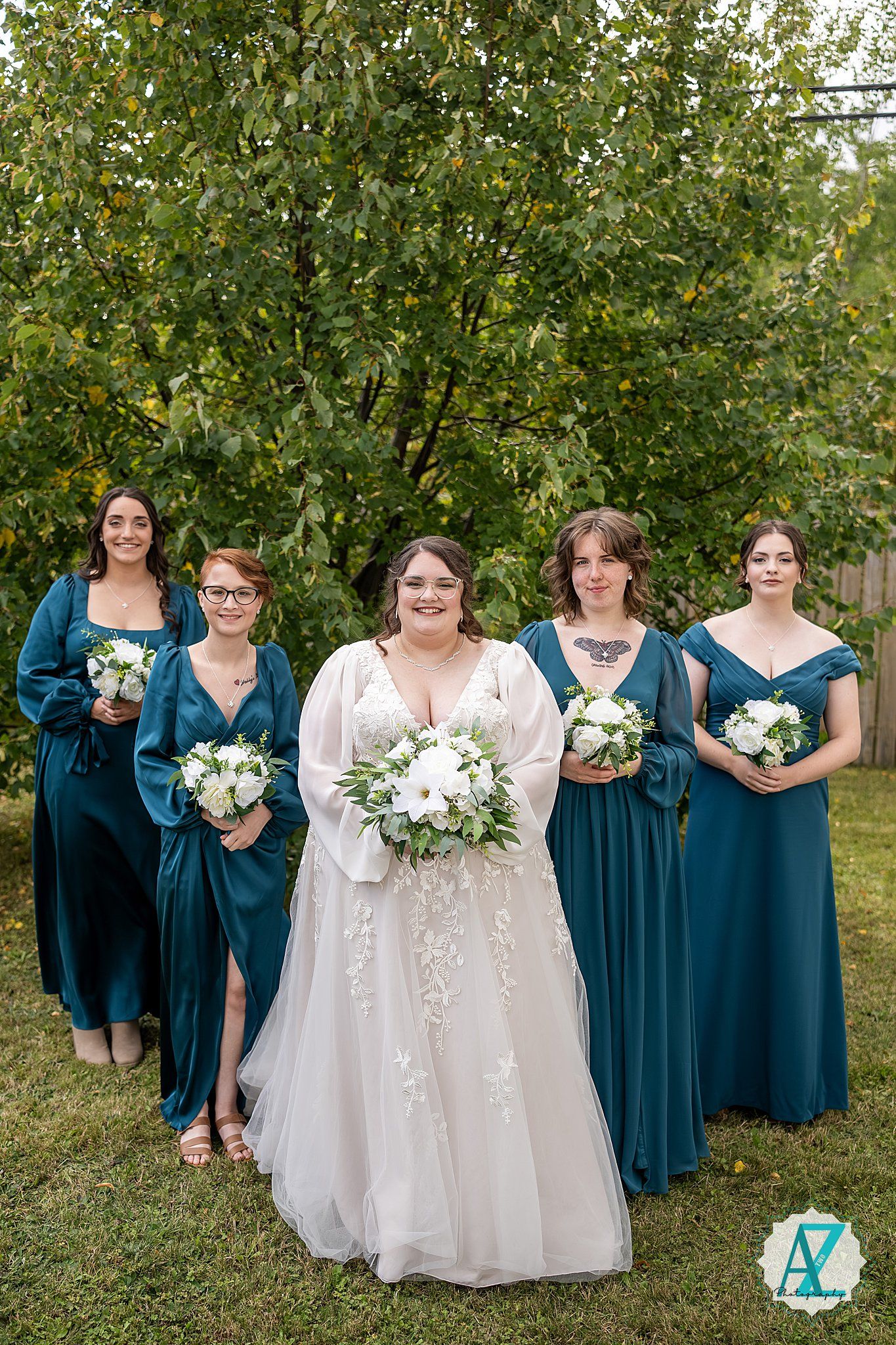 Wedding party members in teal blue and white dresses pose for formal photos in a lush green garden setting.