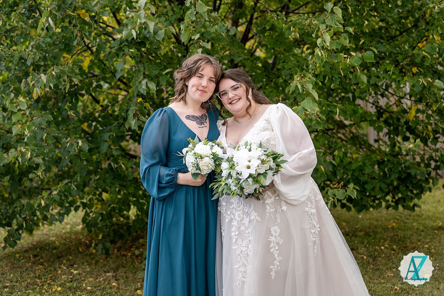 Two people in formal attire stand together holding white floral bouquets against a green leafy background.
