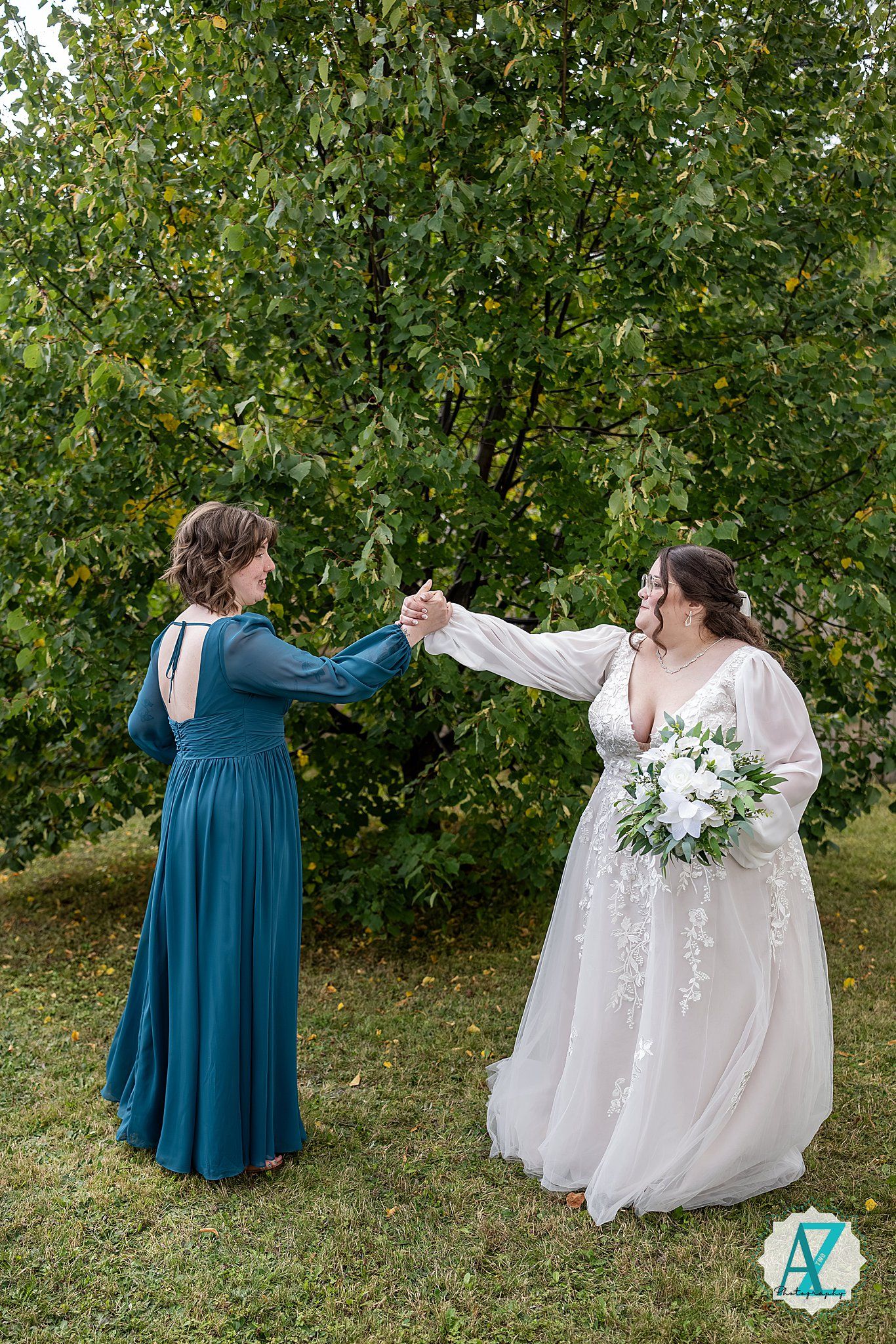 Two people in formal dresses share a joyful moment on a lawn with green trees in the background.