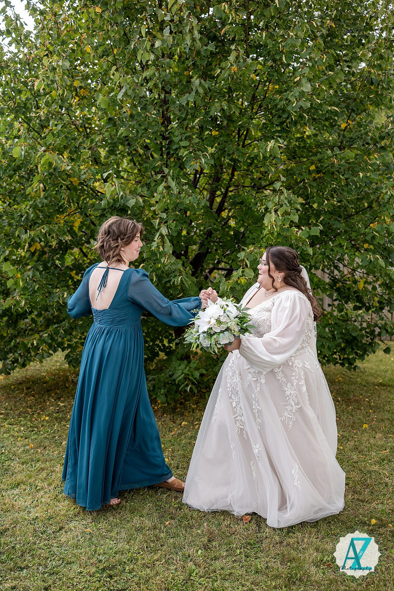 Two people in formal dresses share a joyful moment dancing together on grass with green trees in the background.