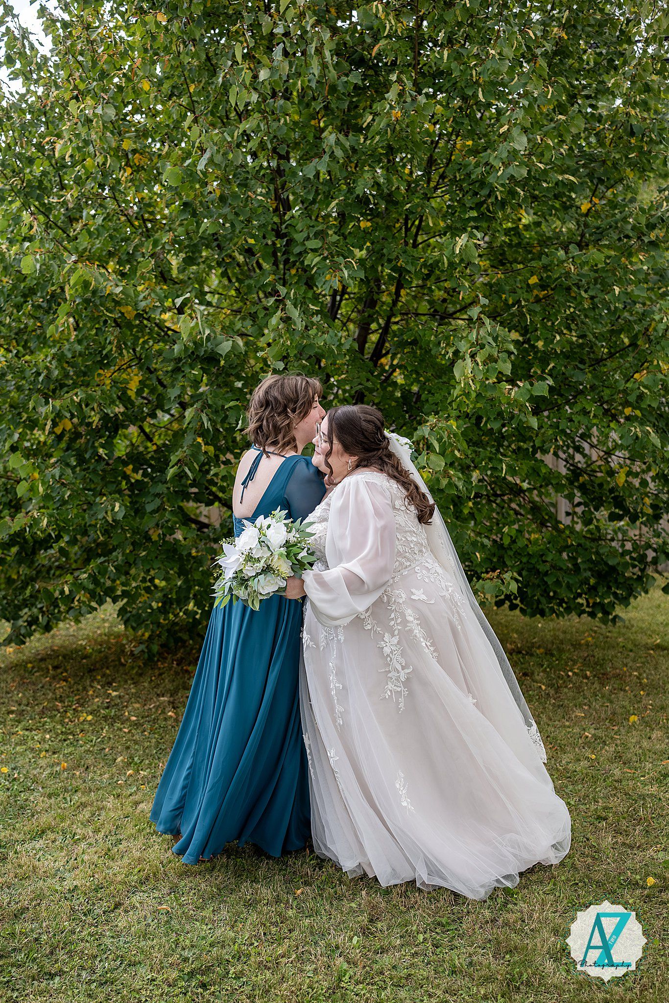 Wedding party members in teal blue dresses and white gown share a joyful moment on a grassy lawn with trees.