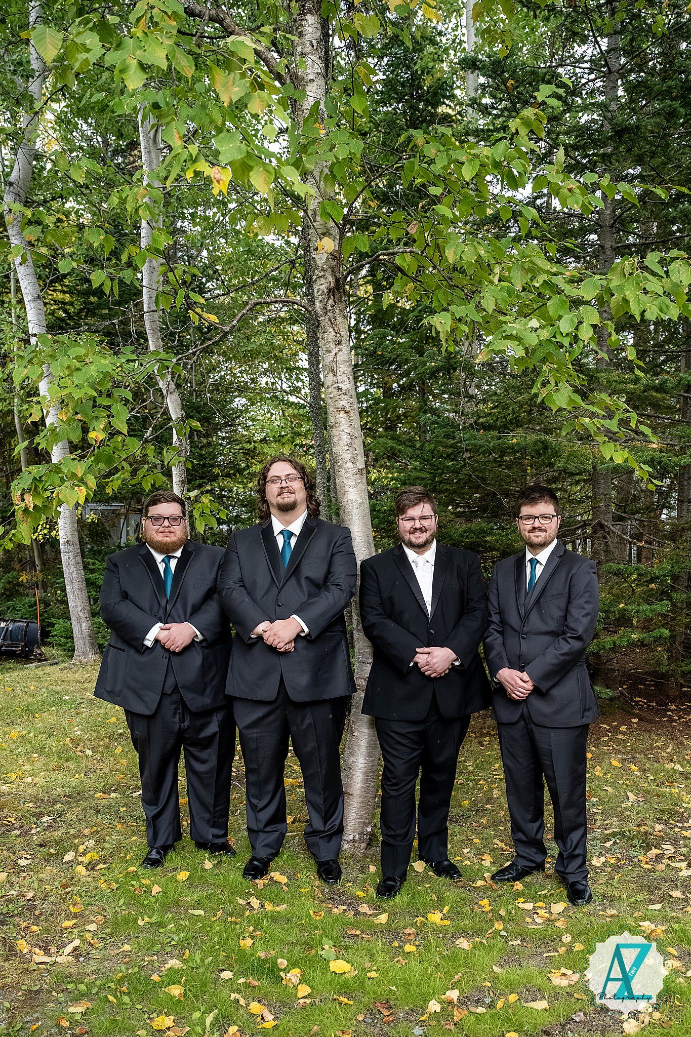 A group of groomsmen in black suits stand together in a wooded outdoor setting with green foliage.