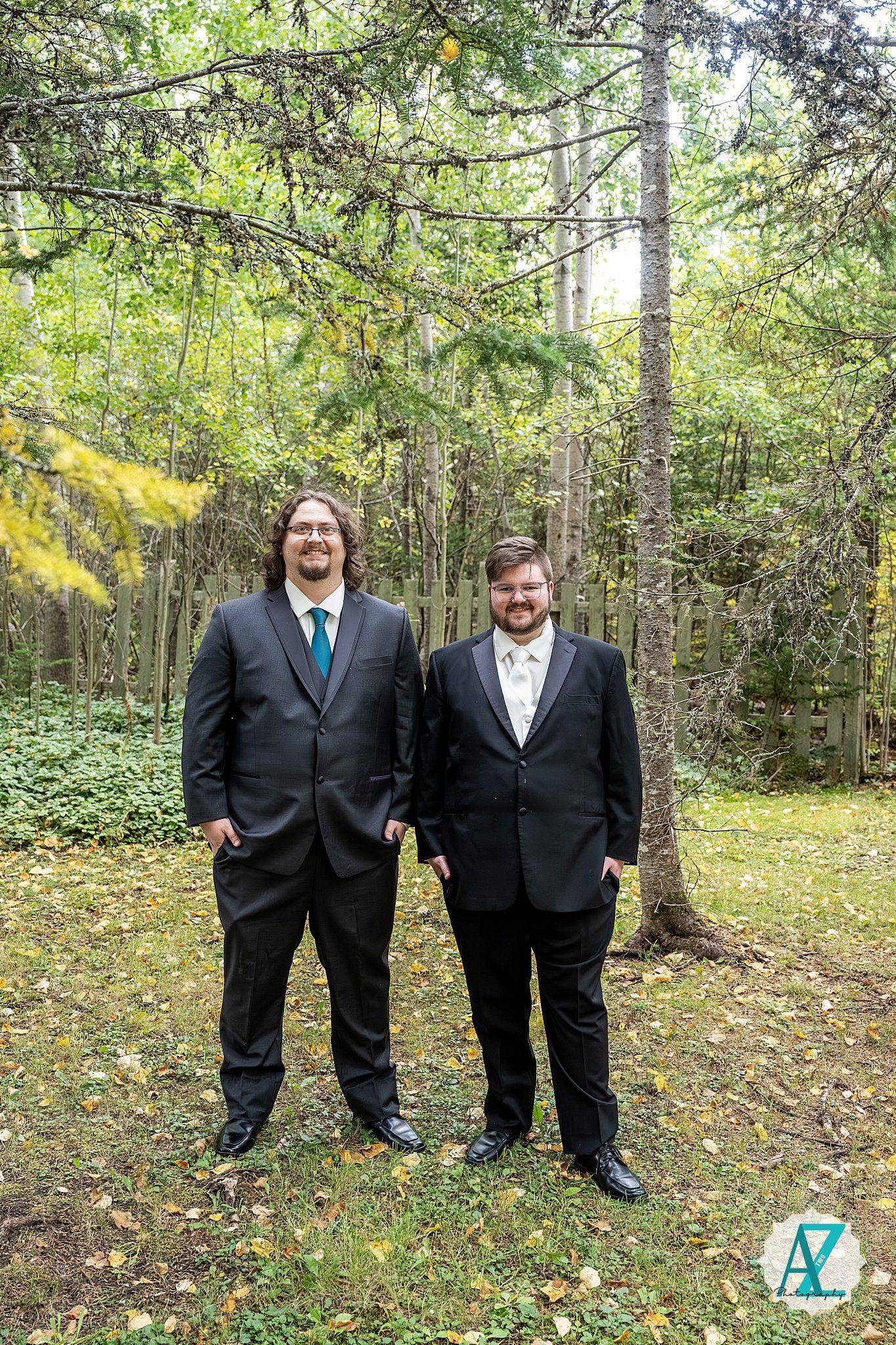 Two people wearing black suits stand together in a wooded outdoor setting with green trees and fallen leaves.