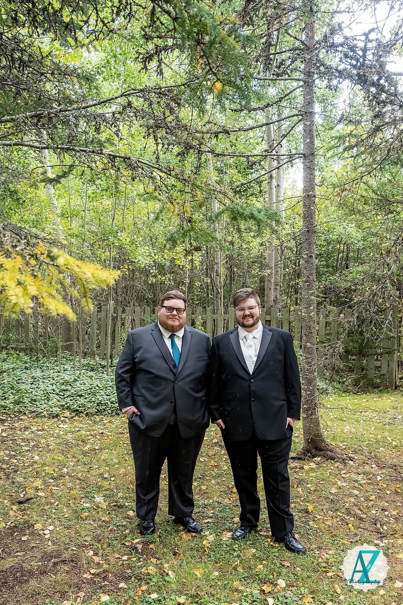 Two people in formal black suits stand together on fallen autumn leaves in a wooded forest setting.