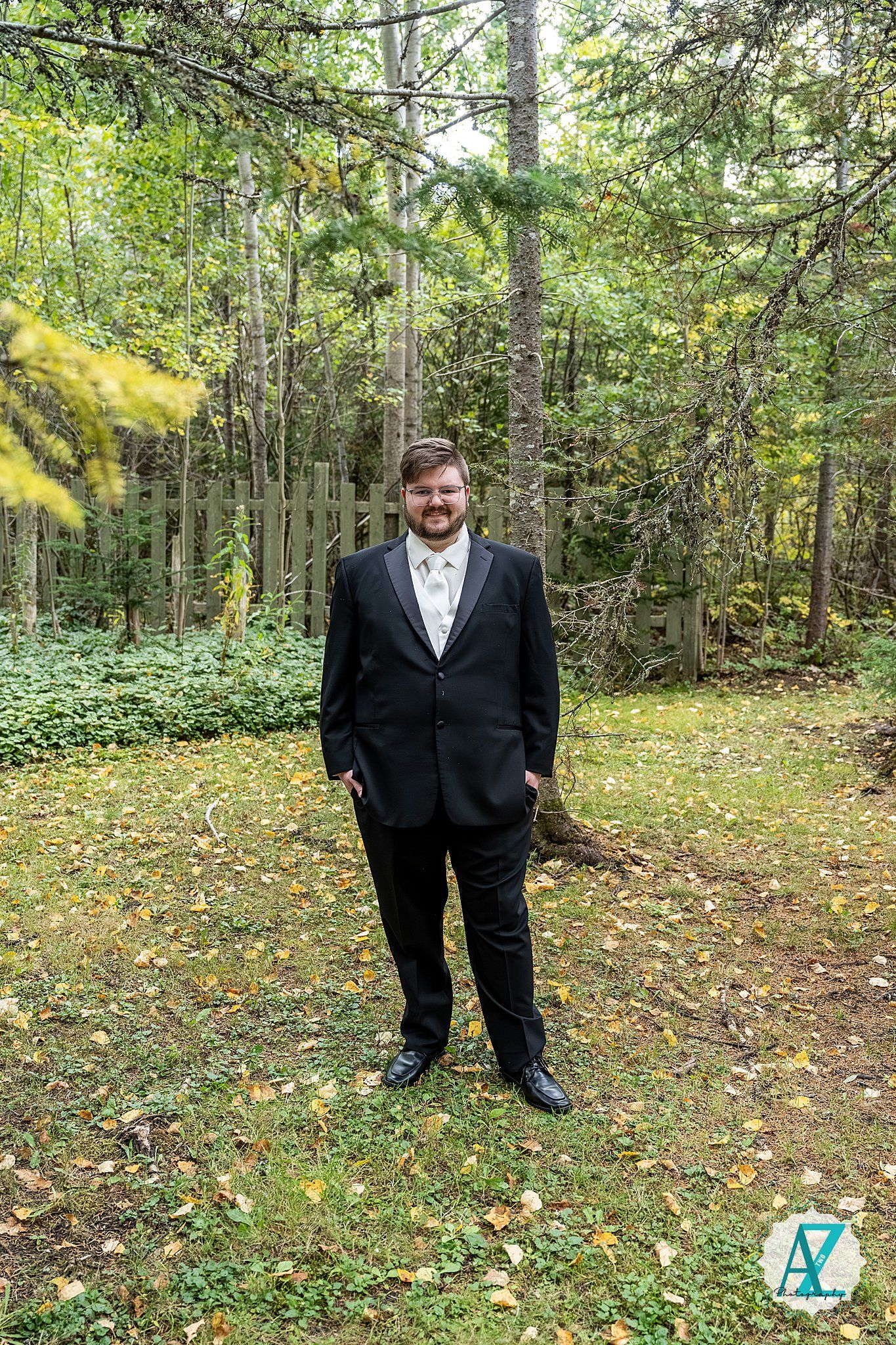 A person wearing a black suit stands on fallen leaves in a wooded outdoor setting.