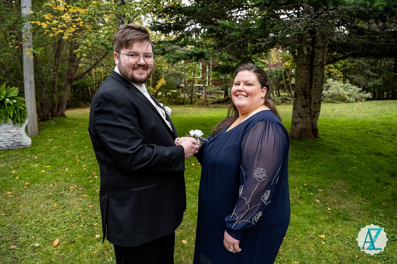 A couple in formal attire stands together on a green lawn with trees in the background during an outdoor event.