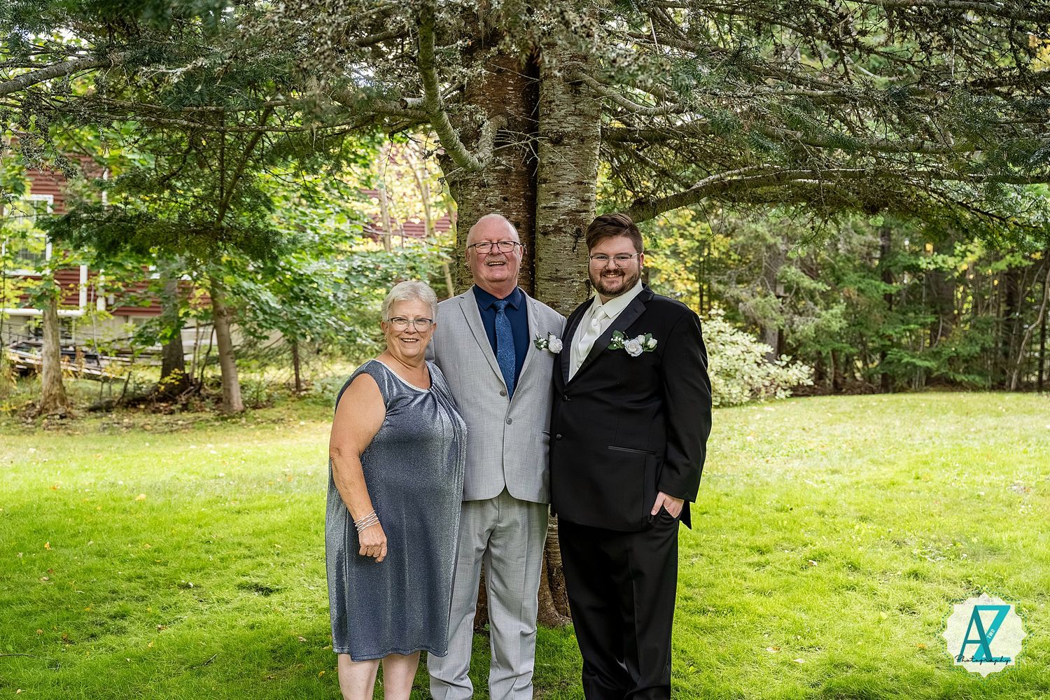 Three people in formal attire pose together on a lush green lawn with evergreen trees in the background.