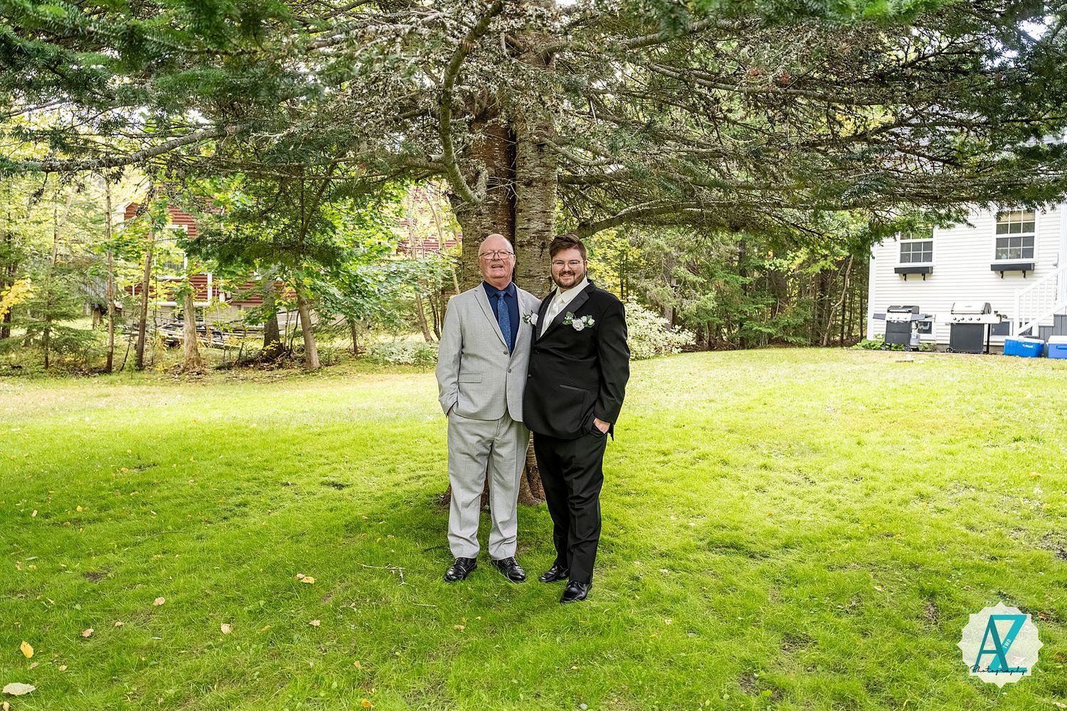 Two people stand together on a green lawn surrounded by tall pine trees on a sunny day.