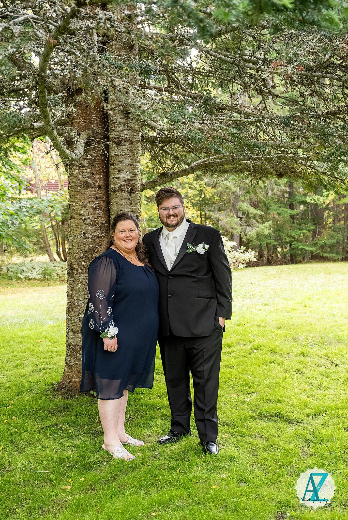 A couple poses together under a large tree on a grassy lawn during a formal event.