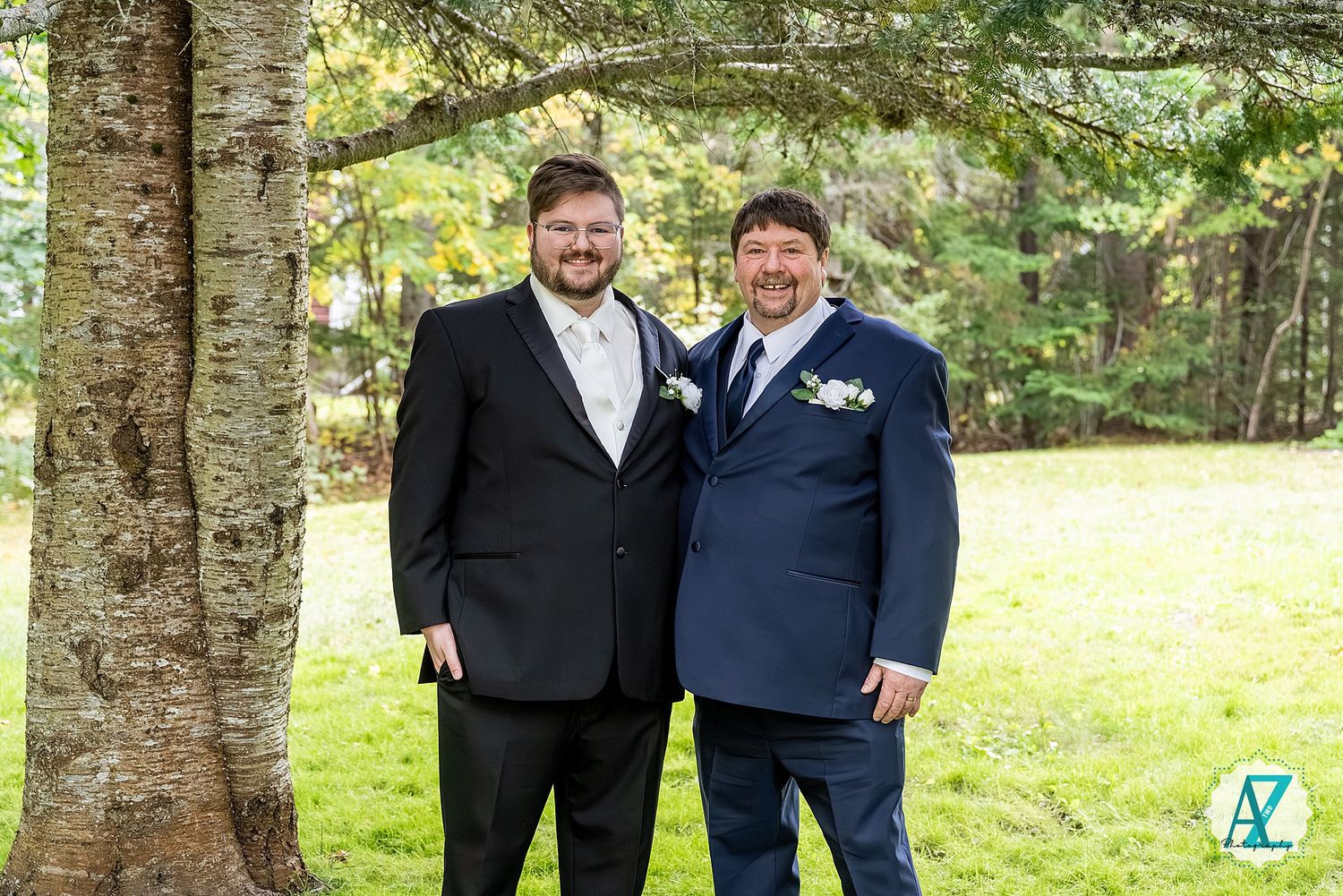 Two people in formal attire stand together outdoors under tall trees in a natural setting.