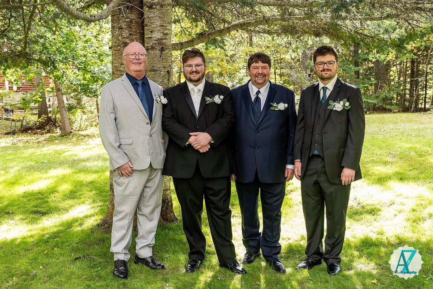 Four people in formal attire stand together on a sunny lawn with evergreen trees in the background.