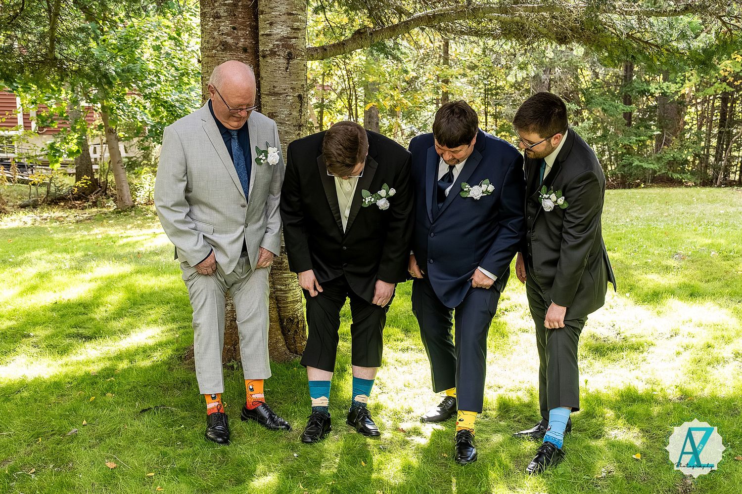 A group of people in formal attire huddle together to examine something on the ground in a sunny outdoor setting.