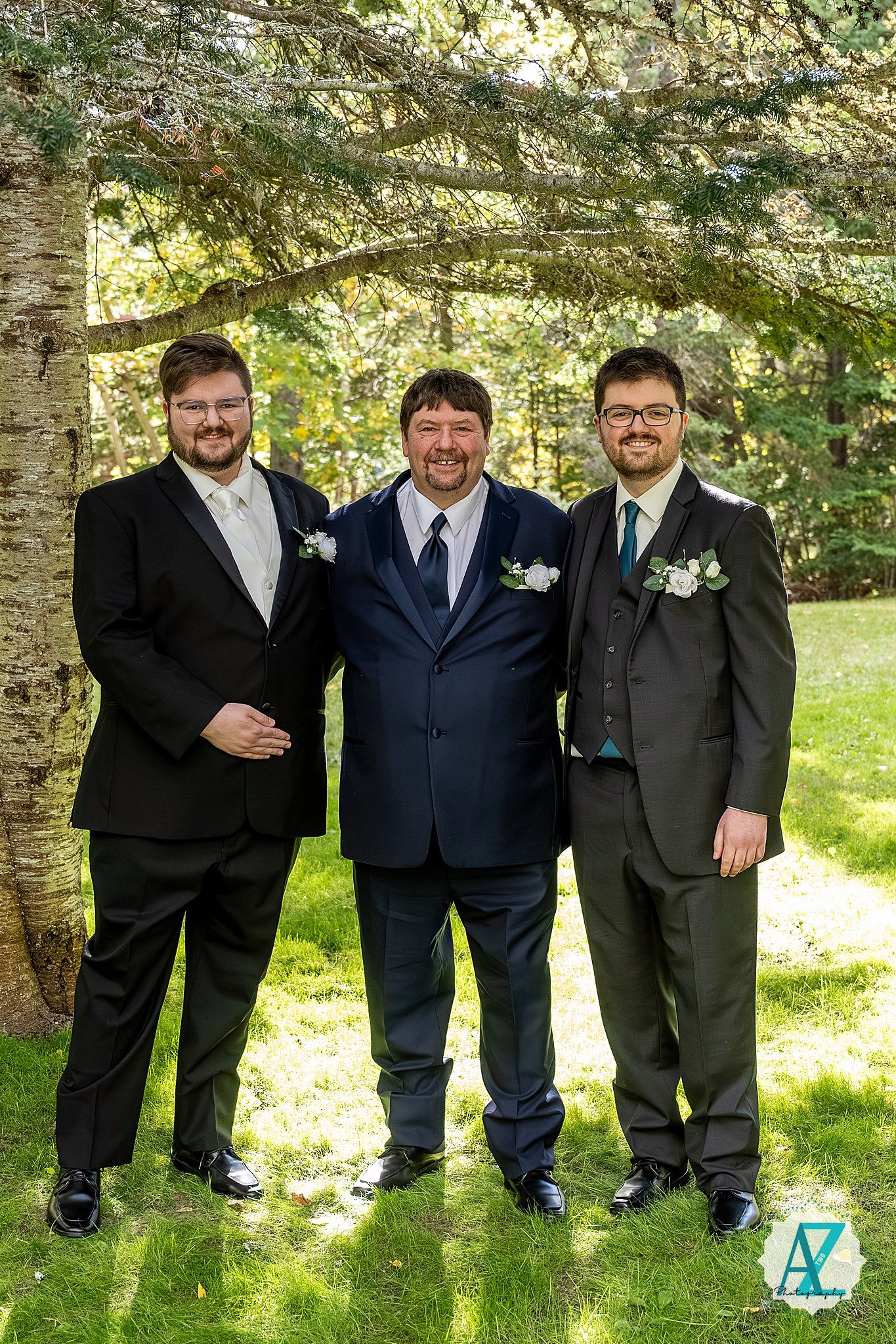 Three groomsmen in formal suits pose together on grass under trees in a natural outdoor setting.
