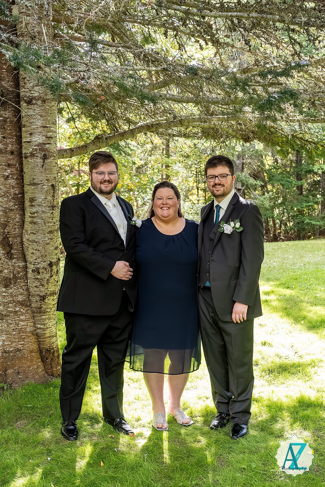 A group stands together in front of evergreen trees on a sunny day wearing formal attire.