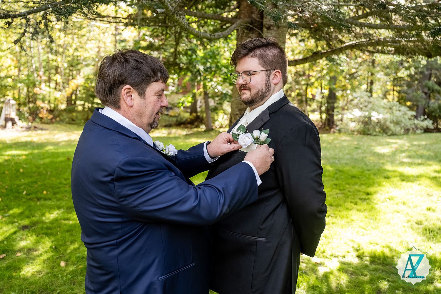 Two people in navy suits share a playful moment while standing outdoors in a sunlit garden setting.