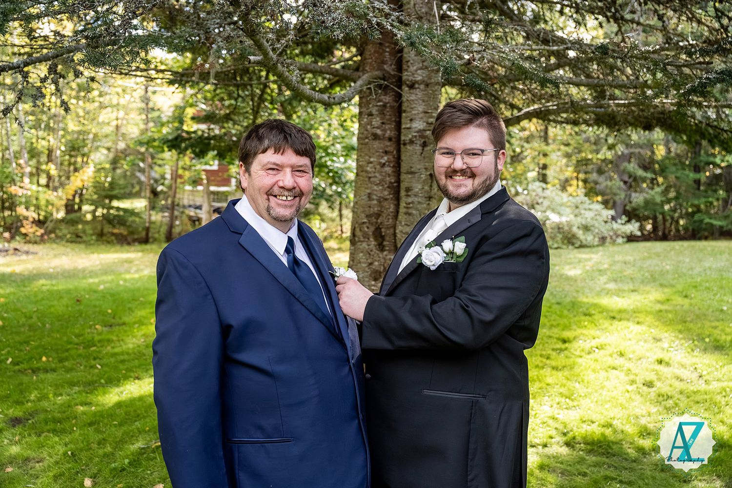 Two people in dark suits share a joyful moment together in an outdoor setting with trees and greenery.