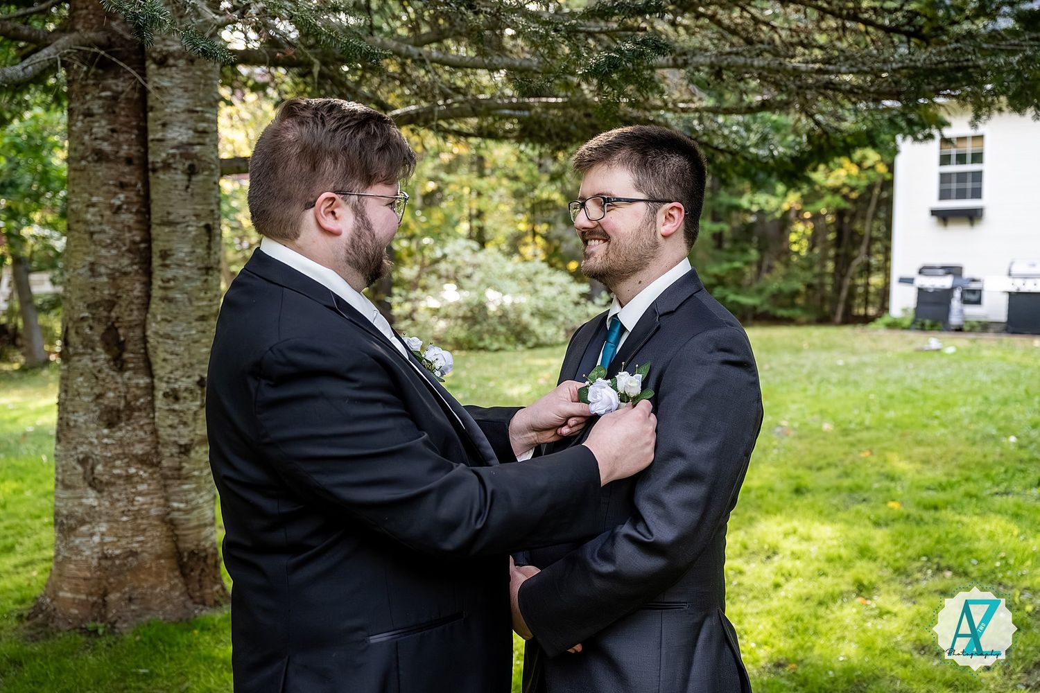 Two people in formal attire laughing together in a wooded outdoor setting with sunlight filtering through trees.