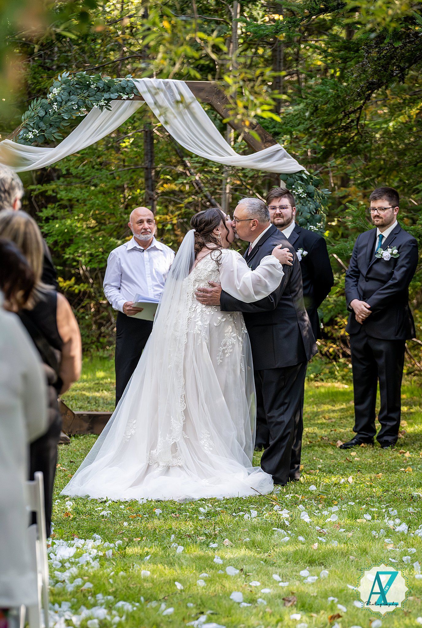 A wedding ceremony takes place outdoors with white draping decor and scattered flower petals on a lush green lawn.