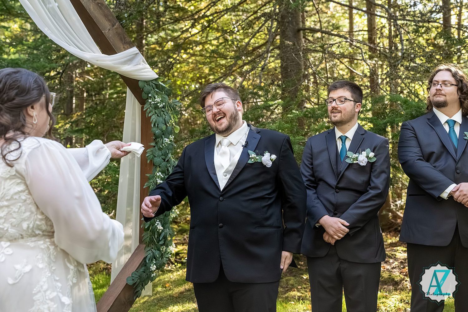 Groomsmen in black suits share a candid laughing moment together in a sunlit wooded outdoor setting.